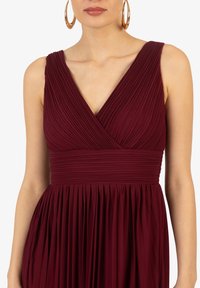 Vinröd plisserad klänning med djup V-ringning, figurnära liv och svävande kjol. Tillverkad av textil som har en mjuk midjeresår.
