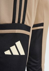 Beige sportbroeken met zwarte accenten, voorzien van drie zwarte strepen aan de zijkant en een groot wit logo op de dij.