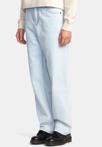 Světle modré, volné džíny s jemným prošíváním, vysokým pasem a předními kapsami, v kombinaci s černými botami na šedém pozadí.