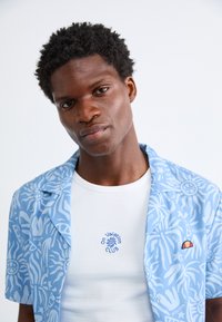 Kortermet, knappet skjorte i lyseblå med hvite abstrakte mønstre, brukt over en tettsittende hvit ribbestrikket tanktop med broderte detaljer.