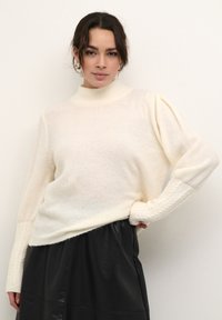 Pull-over crème avec un col montant, des manches texturées et un matériau en tricot doux. Assorti à une jupe en cuir noire.