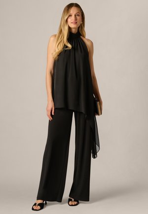 Vrouw met lang blond haar, gekleed in een mouwloos zwarte haltertop en zwarte broek met wijde pijpen, terwijl ze een kleine clutch vasthoudt.