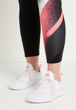 Jambes portant des leggings noirs avec un motif rose et blanc, et des baskets blanches Adidas avec des accents corail sur un sol blanc.