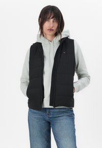 Black padded vest with a fitted design, hood, and zip closure, layered over a light gray hoodie and paired with denim jeans.