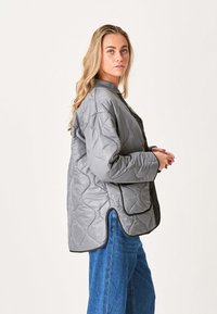 Grå quiltet jakke med afslappet pasform, der har en ribbet krave, lynlås-lommer og kontrasterende sorte accenter langs kanterne.