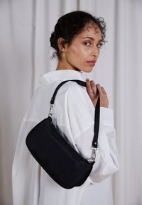 Černá nylonová kabelka s zakřiveným tvarem, se zipovým zapínáním a stříbrnými kovovými doplňky. Nošená přes rameno na bílém tričku.