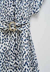 Šaty s leopardím vzorem v bílé barvě, s modrými a černými skvrnami. Tento materiál má texturovaný vzhled, s úzkým pasem a zlatou sponou na pásku.