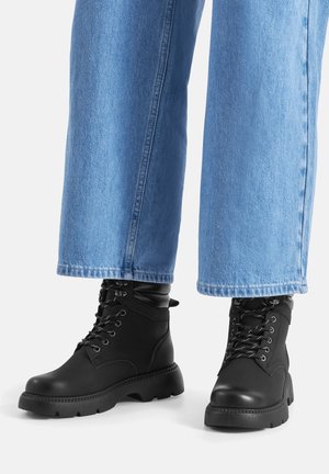 Person, die weit geschnittene hellblaue Jeans und schwarze, Schnürstiefel im Kampfstil trägt, steht auf einer weißen Oberfläche.