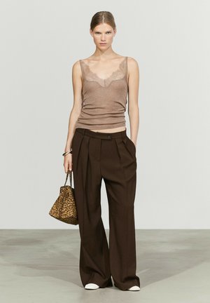 Femme portant un débardeur marron orné de dentelle, un pantalon ample marron foncé, des chaussures blanches, tenant un sac à main imprimé léopard dans un décor de studio neutre.