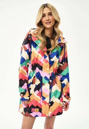 Žena v pestrofarebnom plášti s kapucňou so vzorom cikcak a modrými sťahovacími šnúrkami, stojaca na bielom pozadí.