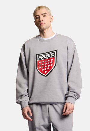 Młody mężczyzna o krótkich blond włosach, ubrany w szarą bluzę i spodnie, bluza ma czerwono-białe logo w kształcie tarczy z napisem "PROSTO."