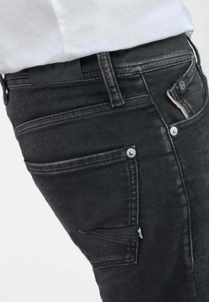 Un jean en denim noir présente une coupe slim, cinq poches et des coutures discrètes. Le matériau montre une légère texture avec un design minimaliste.