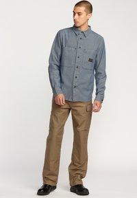 Camisa de botones azul con dos bolsillos en el pecho, combinada con pantalones cargo beige que tienen múltiples bolsillos y zapatos negros.