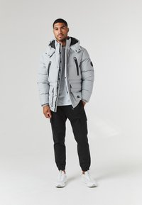 Light gray puffer jacket with a black interior, featuring zippered pockets, logo details, and a hood, paired with black cargo pants and white sneakers.