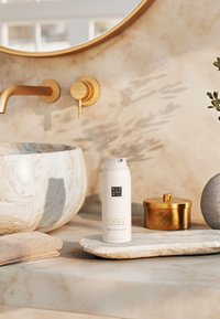 Leave-in ultra curl balm in een witte pompflacon, geplaatst op een licht stenen dienblad naast een gestructureerde handdoek en een decoratieve schaal. Gouden accenten zichtbaar.