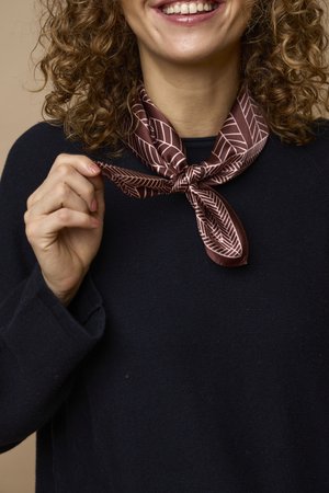 Femme aux cheveux bouclés sourit en tenant l'extrémité d'une écharpe marron à motifs nouée autour de son cou, portant un pull noir.