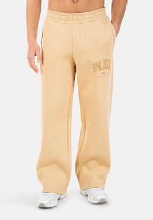 Beige Weitbein-Sweatpants mit elastischem Bund und "SMILODY"-Logo, getragen mit weißen Sportschuhen und Händen in den Taschen.