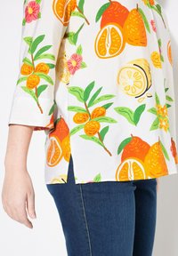 Hvid skjorte med et livligt mønster af appelsiner og citroner, grønne blade og pink blomster. Har 3/4 ærmer og et design med sideslids.