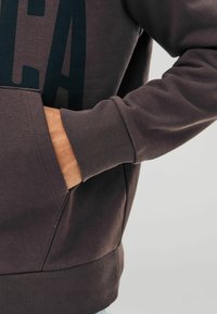 Ruka čiastočne vo vrecku tmavo hnedého hoody s rebrovanými manžetami a pásom, ukazujúca časť veľkých tlačených písmen.