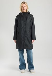 Zwarte, waterdichte regenjas met een capuchon, drukknoopsluiting, zijzakken en een comfortabele pasvorm, gedragen over losse blauwe spijkerbroeken en witte sneakers.
