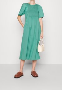 Une robe verte de mi-longueur avec de courtes manches bouffantes et un corsage froncé. Portée avec des sabots bruns et un petit sac à main blanc.