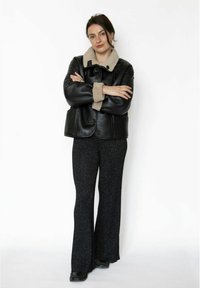 Sort læderjakke med beige pelskrave og manchetter, kombineret med sorte, glitrende vide bukser. Modellen står med armene krydset.