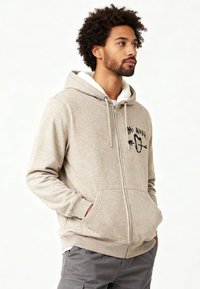 Sweat-shirt zippé beige avec un intérieur blanc en peluche. Il présente un logo sur le devant, des poches latérales et des poignets et ourlet côtelés. Convient pour un usage décontracté.