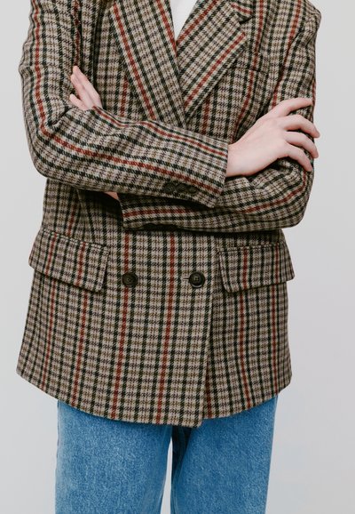 Blazer de corte cruzado en un patrón de cuadro marrón y rojo. Presenta grandes solapas y dos bolsillos delanteros. Se combina con jeans de mezclilla azul claro.