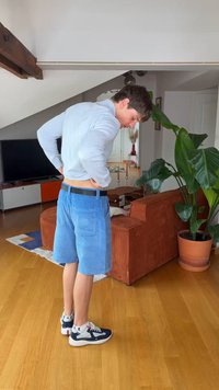Ung mann i blå shorts og lys skjorte ser ned mens han justerer linningen i en koselig stue med en stor plante og oransje sofa.