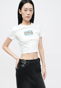 Fehér, rövidített póló, elöl színes grafikával, fekete farmer szoknyával és díszes övvel párosítva.