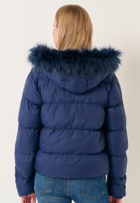 Giacca puffer blu navy con cappuccio in finto pelliccia scura e morbida. Presenta un'imbottitura orizzontale, una silhouette aderente e polsini elasticizzati.