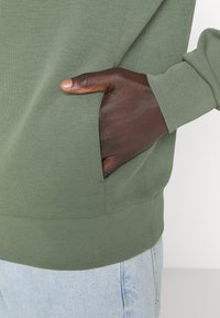 Zelená mikina s žebrovanou texturou, s boční kapsou a uvolněným střihem. Spárována se světle modrými džíny.