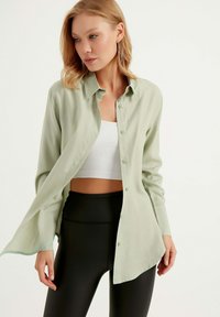 Camicia button-up verde chiaro realizzata in tessuto setoso, con una vestibilità comoda, maniche lunghe e orlo curvo. Indossata sopra un top corto bianco e leggings neri.