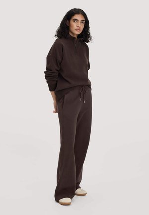 Person mit lockigem dunklem Haar, die einen dunkelbraunen Sweatshirt mit Reißverschluss und passende lockere Hose trägt, dazu cremefarbene Schuhe auf neutralem Hintergrund.