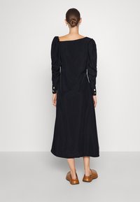 Svart, långärmad klänning med fyrkantig neckline, puffärmar och manschettdetaljer. Slät textur, knälång längd, bärs med bruna tofflor.