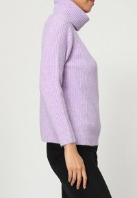 Pull à col roulé lilas en côte avec des manches longues, texture douce et coupe décontractée, associé à un pantalon noir.