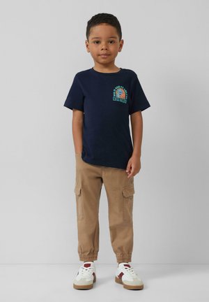Marineblaues Baumwoll-T-Shirt mit einem Löwengrafik; beige Cargo-Hosen mit Seitentaschen; weiße Sneaker mit bordeauxroten Akzenten; lockere Passform.