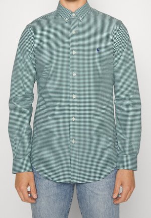 Mann trägt grünes und weiß kariertes langärmeliges Hemd mit kleinen gesticktem Logo auf der linken Brust und hellblaue Jeans.