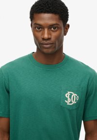 Camiseta de algodón verde con mangas cortas, cuello redondo y un bolsillo en el pecho izquierdo que presenta un diseño de logo en crema. Tiene un acabado texturizado.