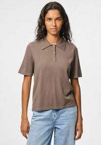 Brun kortärmad polo med platt krage och knäppning med tre knappar, tillverkad av strukturerad tyg, ihop med ljusblå jeans.