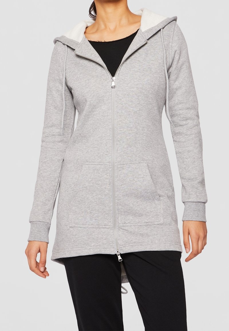 Sweat à capuche gris zippé, doublé en polaire au niveau de la capuche, deux poches frontales, manches longues avec ourlets aux poignets, et un ourlet arrière légèrement plus long.