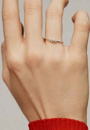 Anillo metálico delgado en oro con tres piedras preciosas claras agrupadas en una mano; el fondo es neutral con una apariencia de piel texturizada.