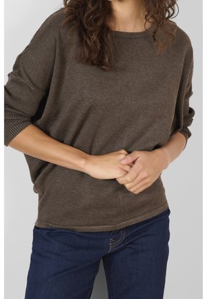 Femme portant un pull marron à manches longues et un jean bleu, debout les mains jointes à la taille devant un fond gris uni.