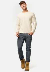 Beige gestricktes Langarmshirt, graue Jeans und braune Stiefel. Das Shirt hat einen runden Halsausschnitt und ein strukturiertes Material.