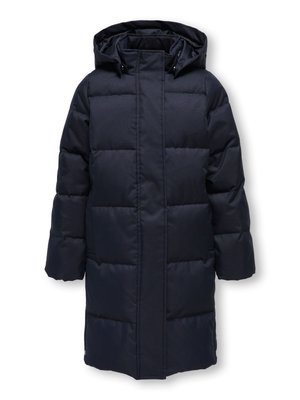 Barn med långt vågigt hår som bär en lång svart vadderad jacka, randig tröja, blå jeans och vita sneakers, ler med händerna i fickorna.