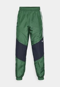 Groene en marineblauwe windbreaker-broeken met een elastische tailleband, zijstrepen en een gladde, glanzende textuur met schuine kleurvlakken.