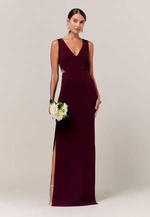 Ženska v brezrokavu globoko vijolični obleki z V-izrezom, ki drži bele cvetove, dopolnjena z zlatimi uhani in zapestnico.