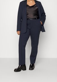 Marineblauer maßgeschneiderter Anzug mit strukturiertem Blazer und geraden Hosen, kombiniert mit einem schwarzen Satin-Camisole und klobigen schwarzen Slippern.