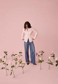 Frau in pink gestreiftem Hemd und weitem Jeans steht zwischen Glasvasen mit pinken Rosen auf einem pinken Boden und Hintergrund.