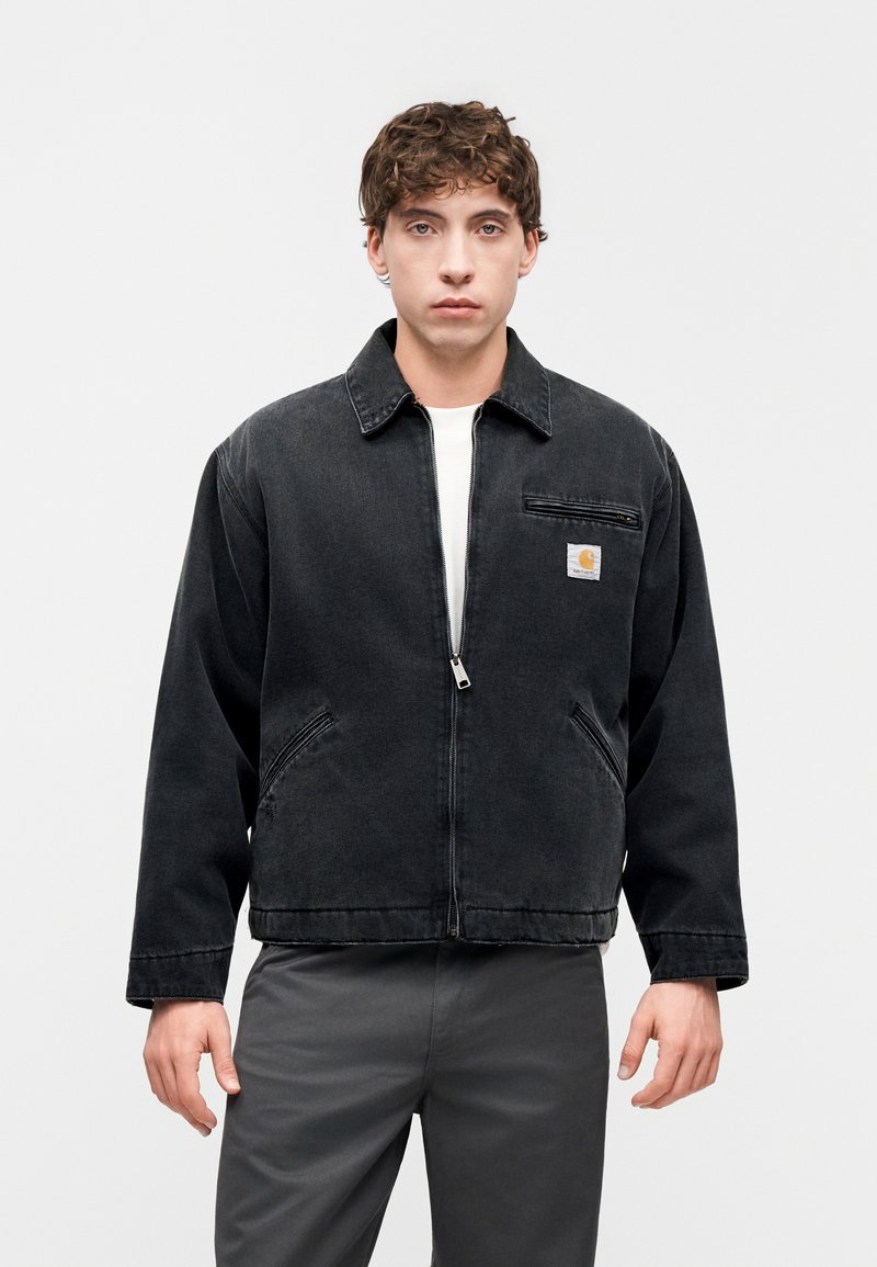 Giacca di jeans nera con zip frontale, colletto a punta e tasche laterali. Presenta una piccola patch con logo sulla tasca del petto. Tessuto texturizzato.